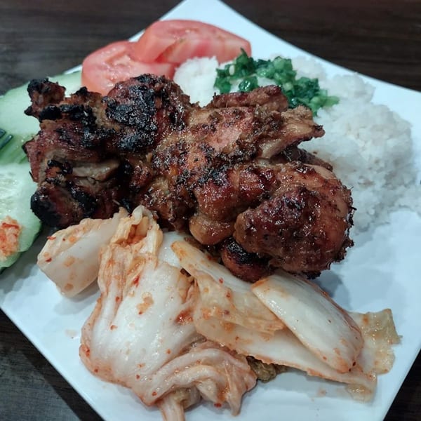 Grilled chicken with rice, sliced tomatoes, cucumbers, and kimchi on a white plate.