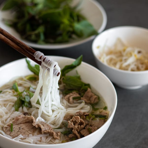 Chopsticks lift pho noodles from a bowl with beef, herbs, and broth, with side dishes.