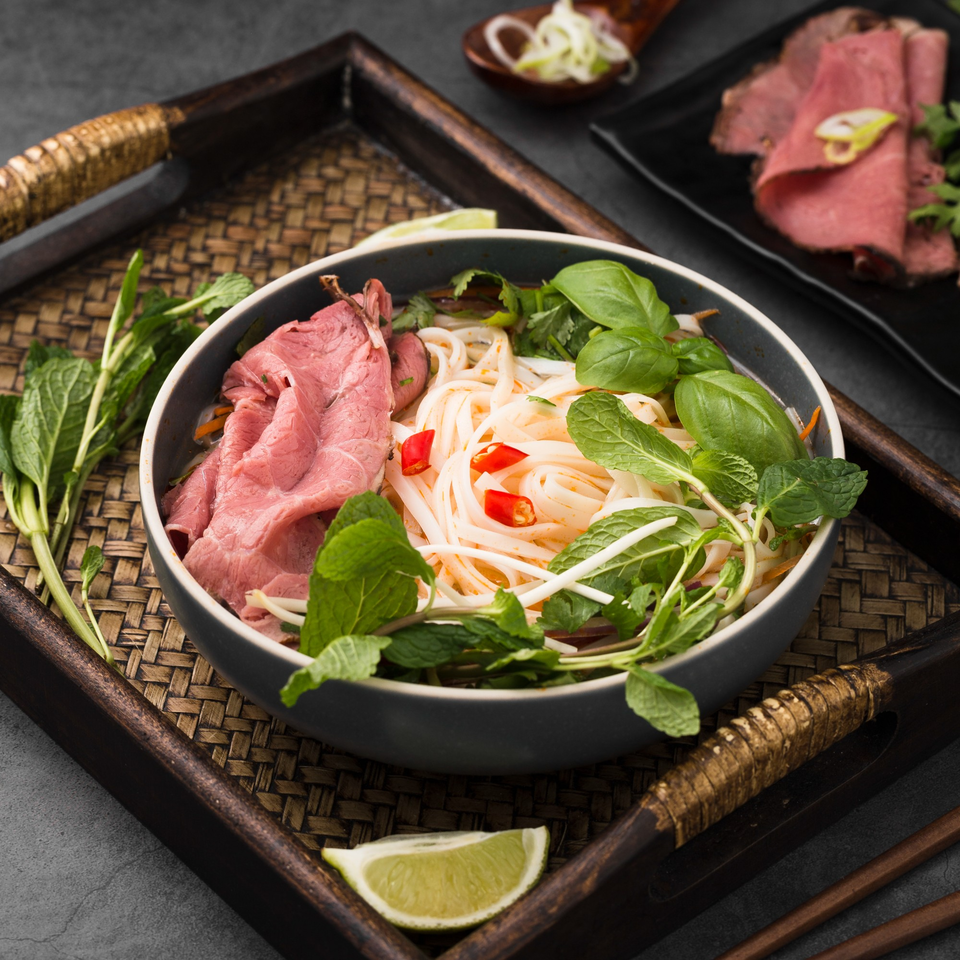 A bowl of Pho 9 pho with thinly sliced beef, noodles, herbs, and chili on a woven tray.